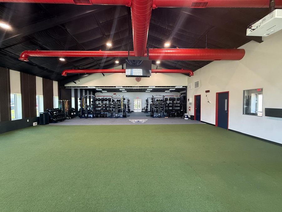 The turf hall on axis: red ductwork crossing a black ceiling, the weight room framed in the opening beyond